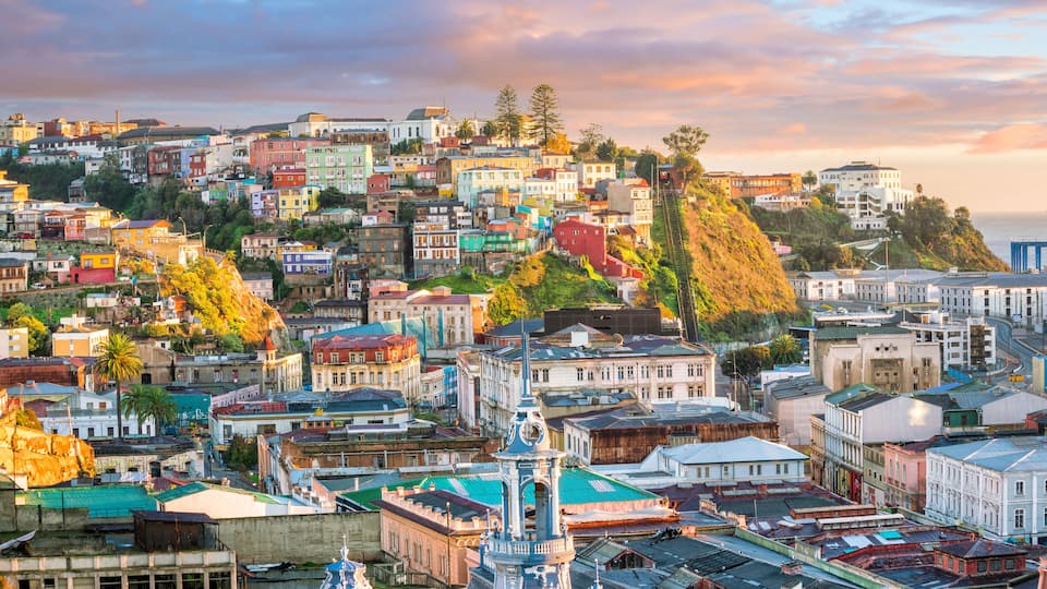 A densely packed urban landscape with colorful buildings, a prominent church spire, and a hillside with trees.