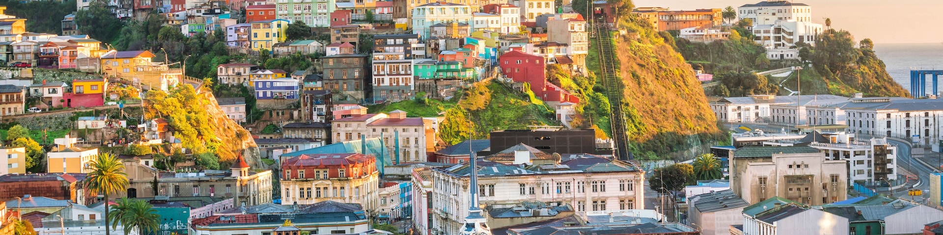 A densely packed urban landscape with colorful buildings, a prominent church spire, and a hillside with trees.