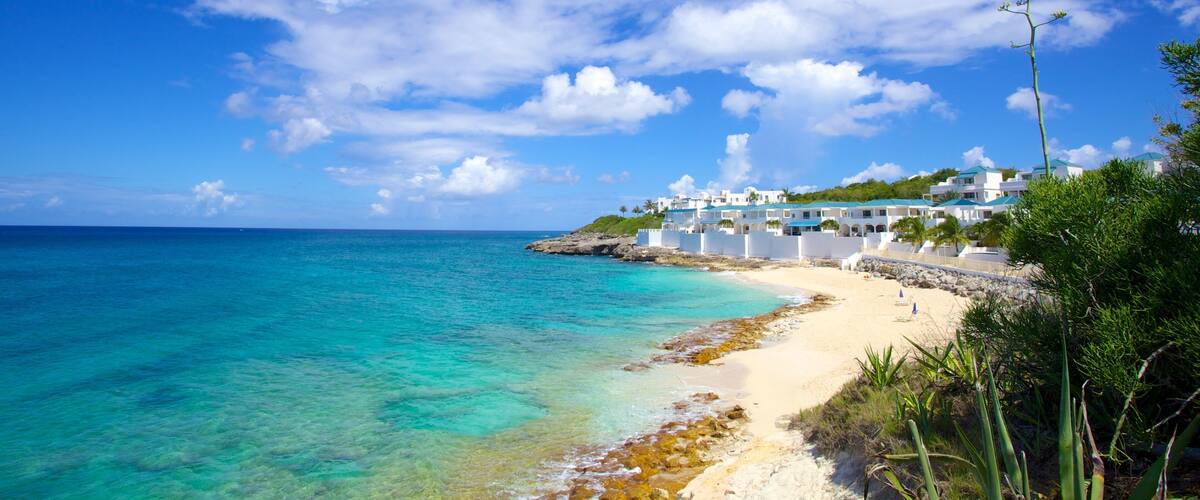 A coastal beach with clear turquoise waters, white buildings, and a rocky shoreline.