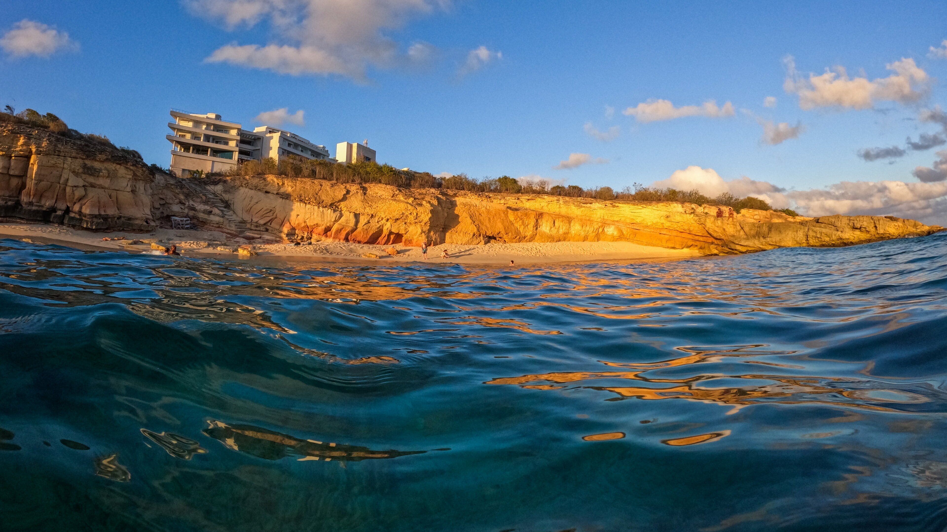 A coastal scene with clear blue waters, a sandy beach, rocky cliffs, and modern buildings on the cliff.