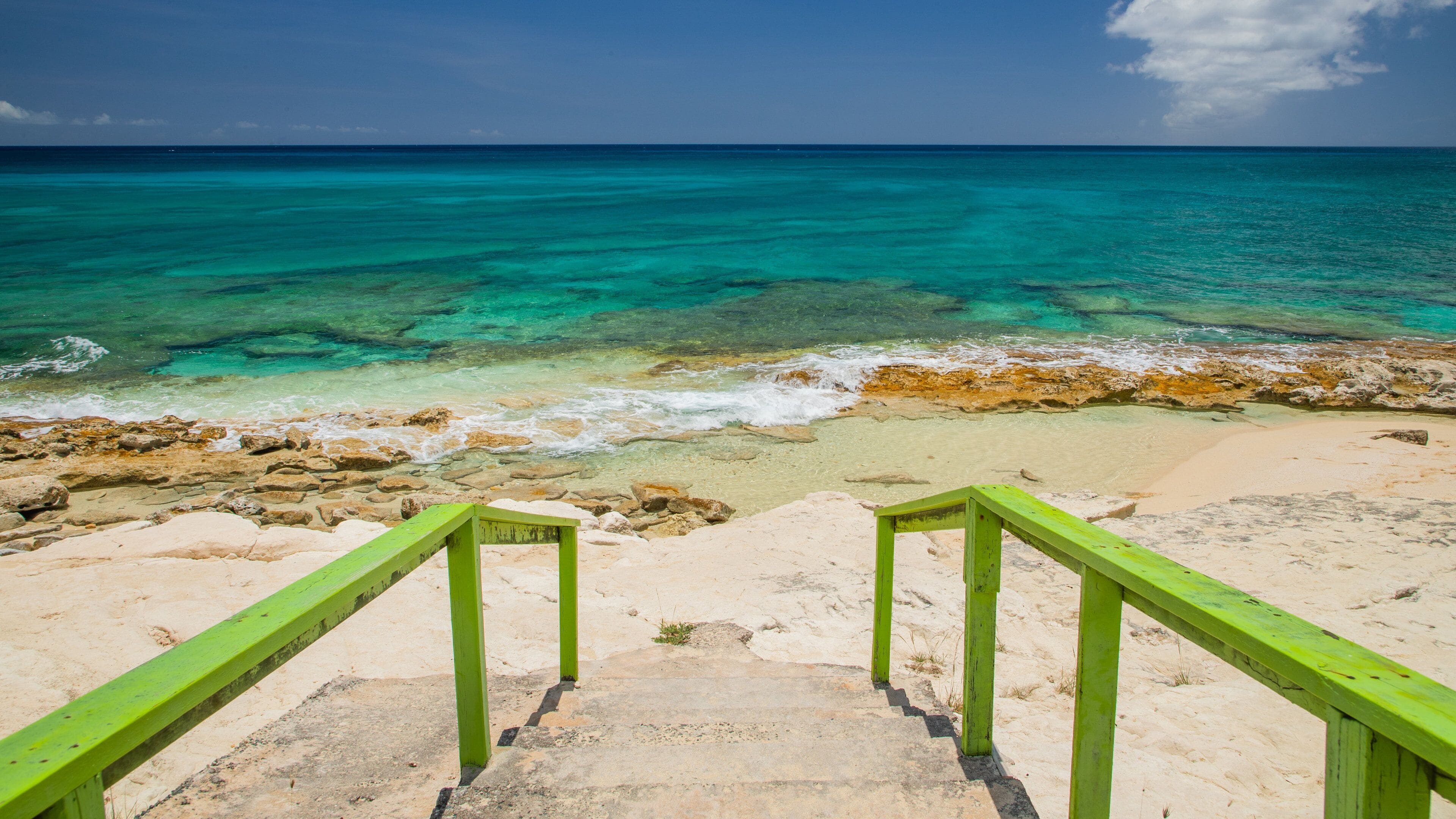 A beach with turquoise waters, a sandy shore, and green stairs leading to the water.
