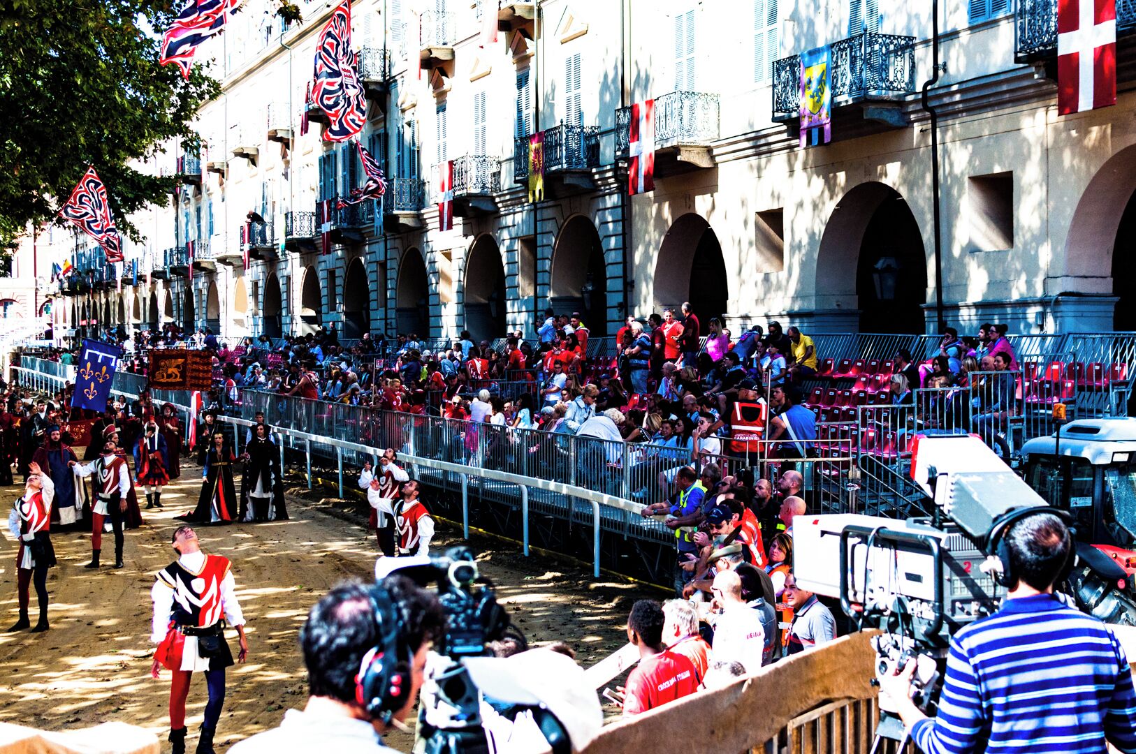 A historical reenactment with participants in period costumes, a crowd of spectators, and a backdrop of classical architecture.