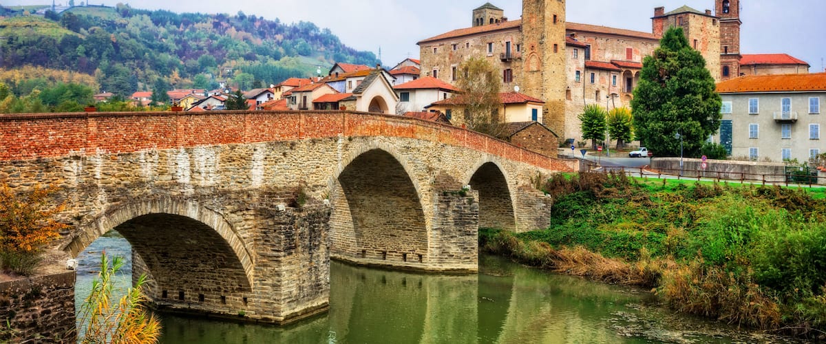 A stone bridge over a river, a historic building with a tower, and a hillside with trees.