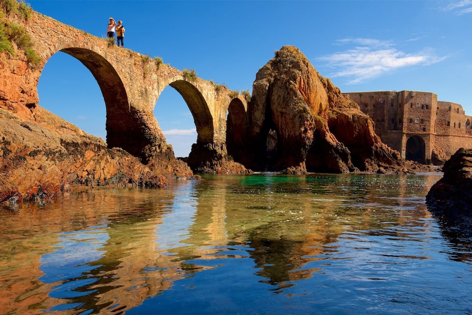 A stone bridge over a river, with rocky cliffs and a fort in the background.