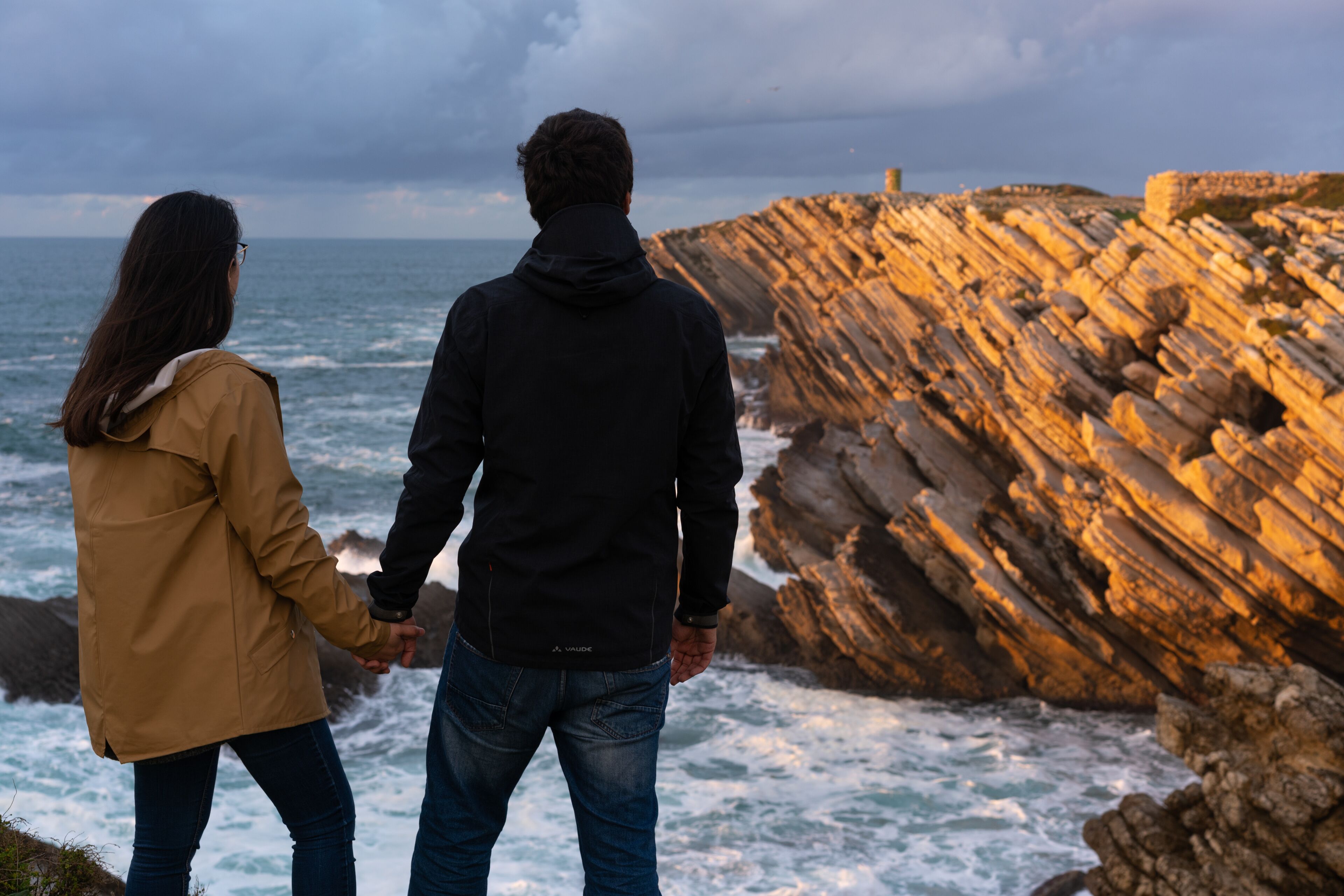 Una pareja tomándose de las manos junto a una costa rocosa con olas rompiendo contra las rocas.