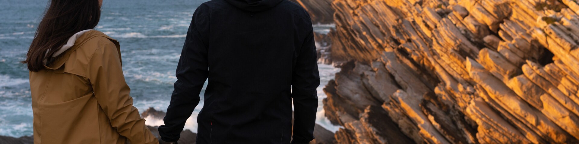 Una pareja tomándose de las manos junto a una costa rocosa con olas rompiendo contra las rocas.