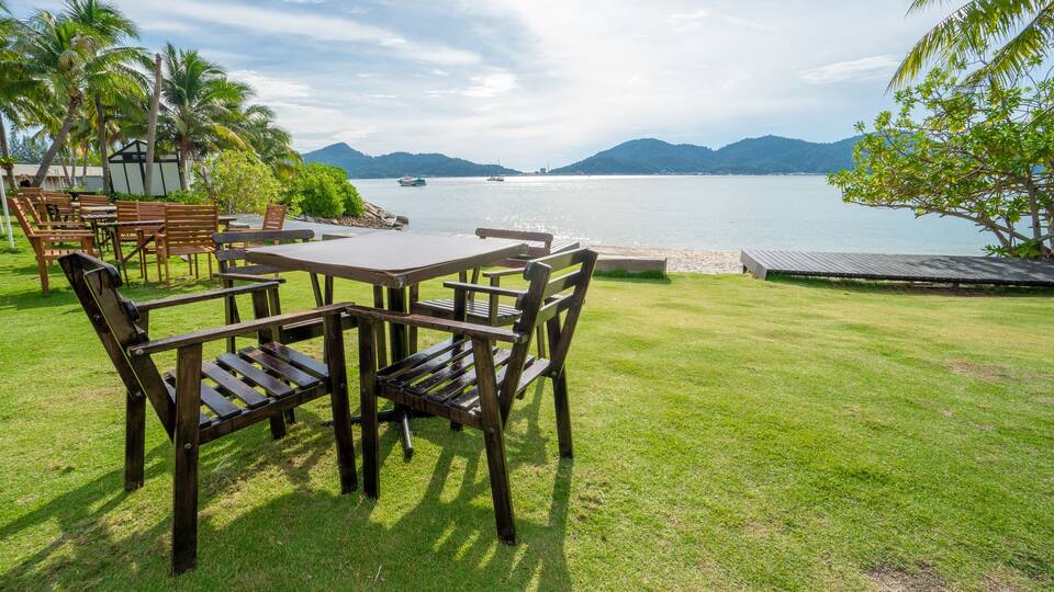 A grassy area with wooden tables and chairs, a wooden deck, and a view of a calm body of water with boats and mountains in the distance.
