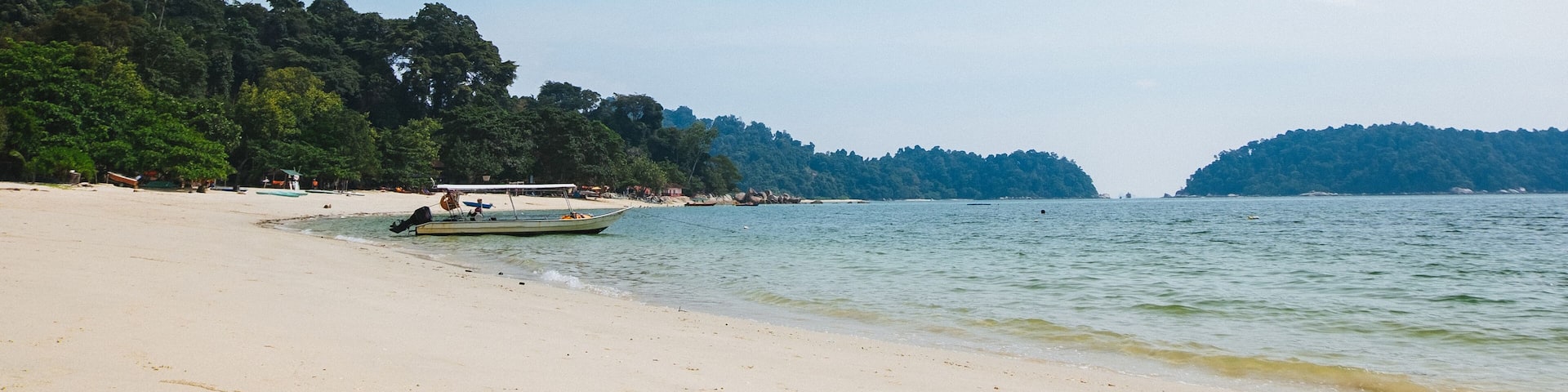 A beach with a boat, calm water, and forested hills in the background.