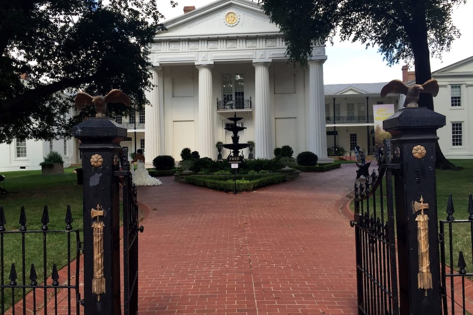 Una entrada imponente con un camino de ladrillo que lleva a un edificio blanco con columnas y un reloj.