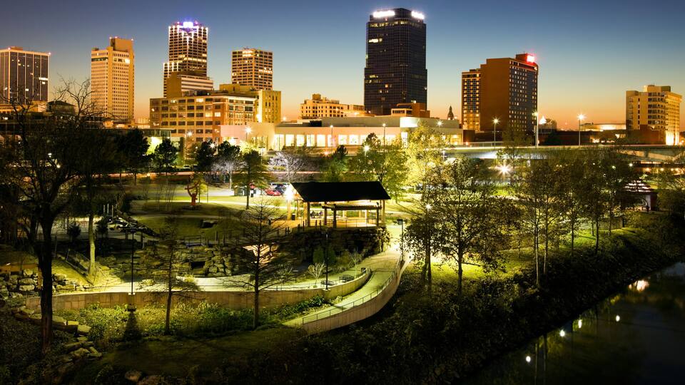 A city skyline at dusk with illuminated buildings, a park with pathways, and a body of water reflecting lights.
