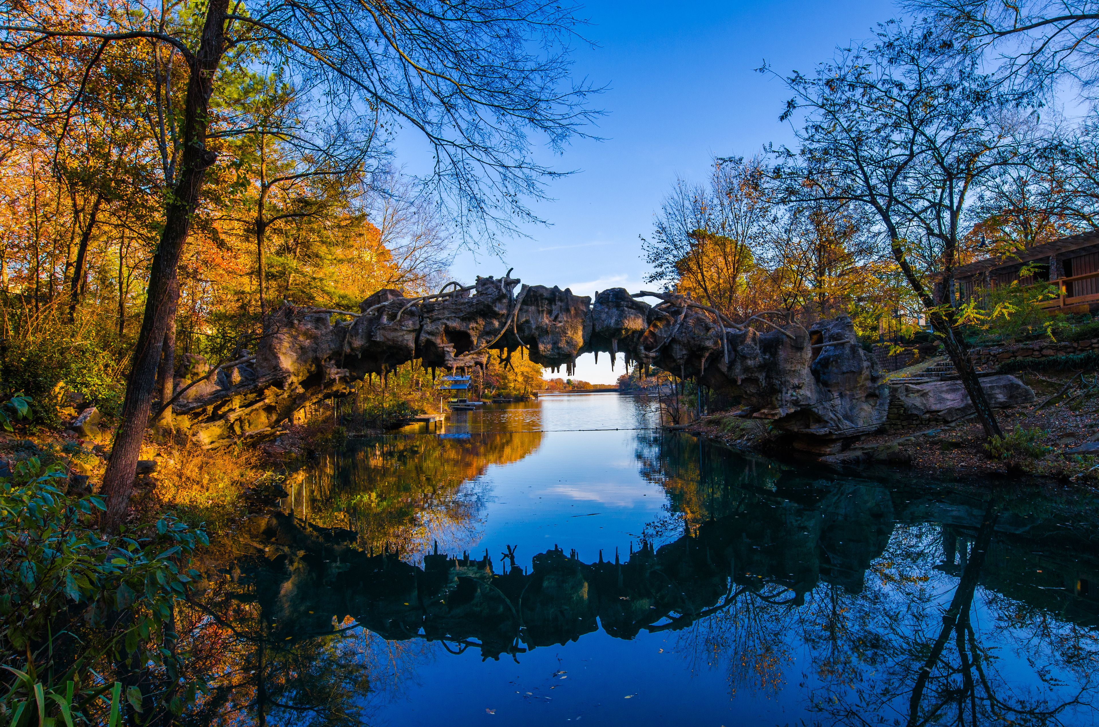 A serene river with a rocky bridge, surrounded by autumn-colored trees and clear blue sky.