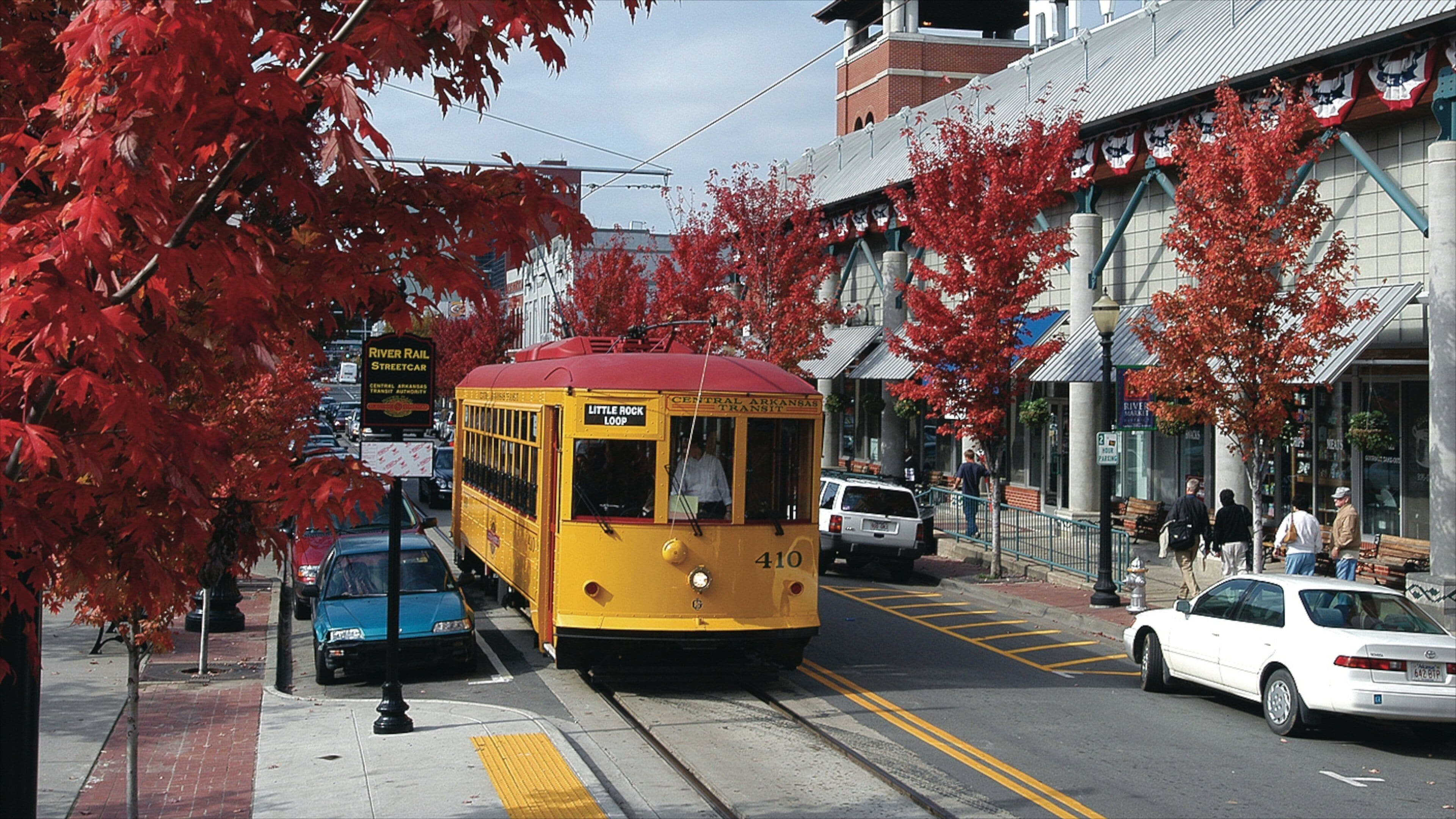 Een gele trolley met de tekst 'Little Rock Loop' op een straat met rode herfstbladeren, auto's en voetgangers.