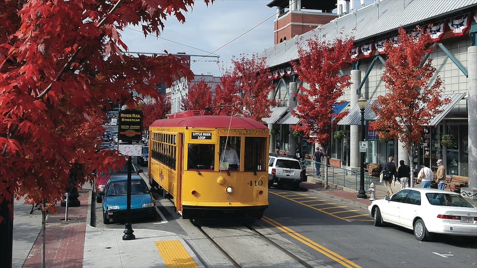 Een gele trolley met de tekst 'Little Rock Loop' op een straat met rode herfstbladeren, auto's en voetgangers.