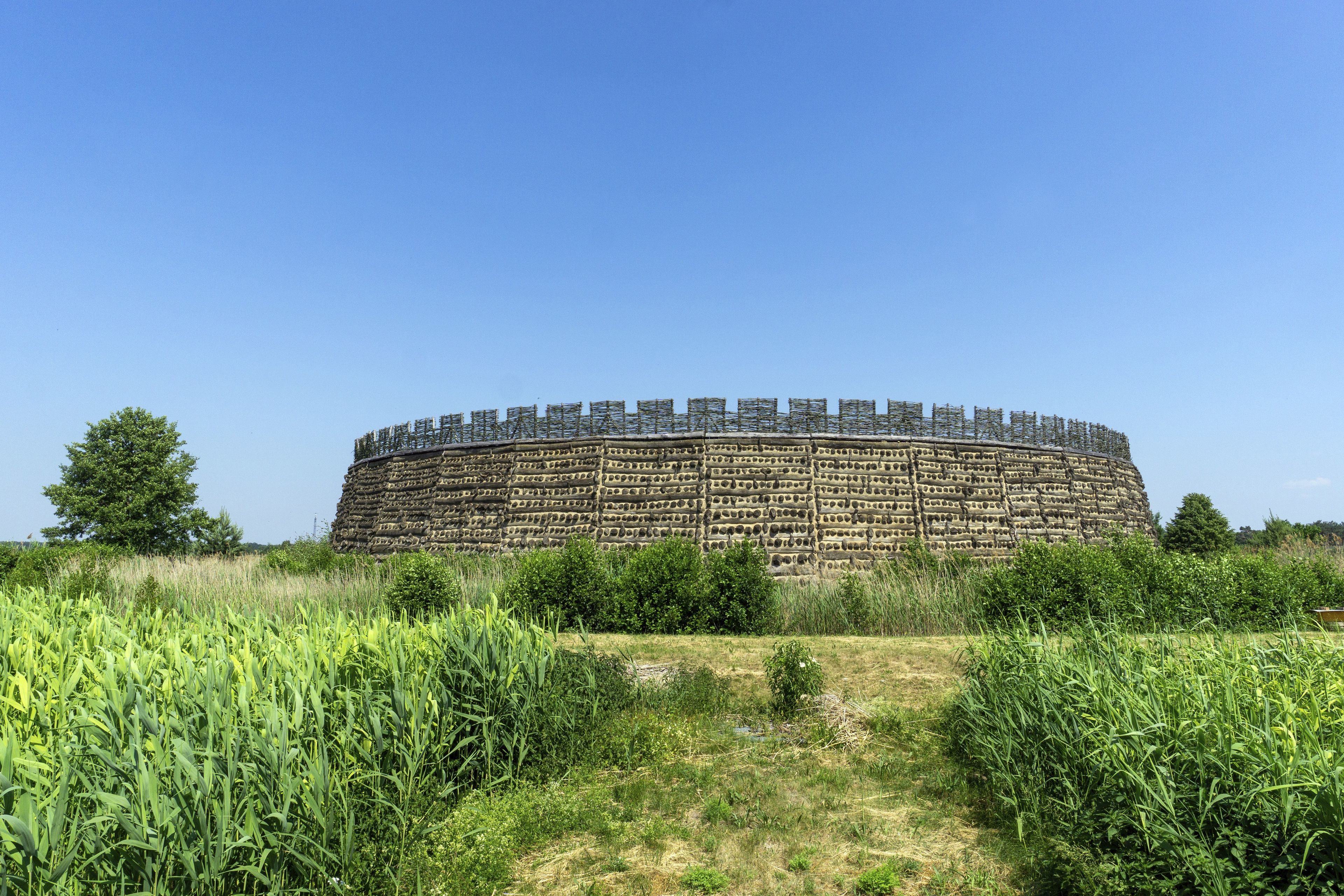 Gran estructura de pedra circular amb una superfície plana, envoltada per un prat d'herba alta i alguns arbres.