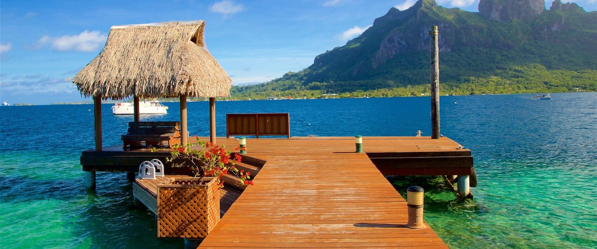 A wooden pier extending into clear turquoise waters, a thatched-roof structure, a bench, and a mountain in the background.