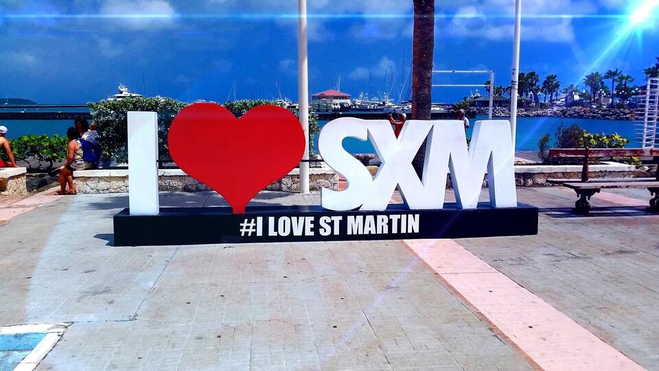 A sign with 'I ♥ SXM' and '#1 LOVE ST MARTIN' in a public space with a view of a marina and boats.