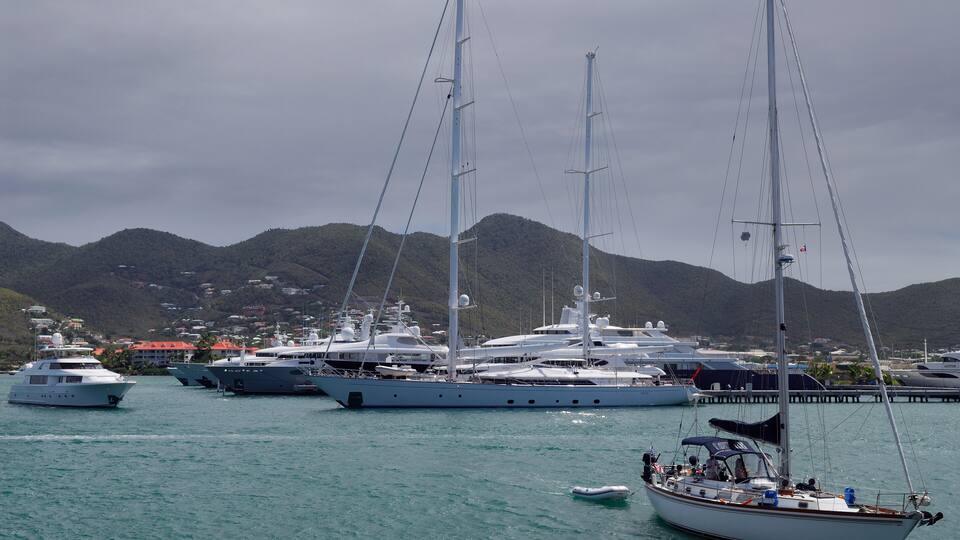 A marina with several sailboats and yachts docked, surrounded by hills and a cloudy sky.