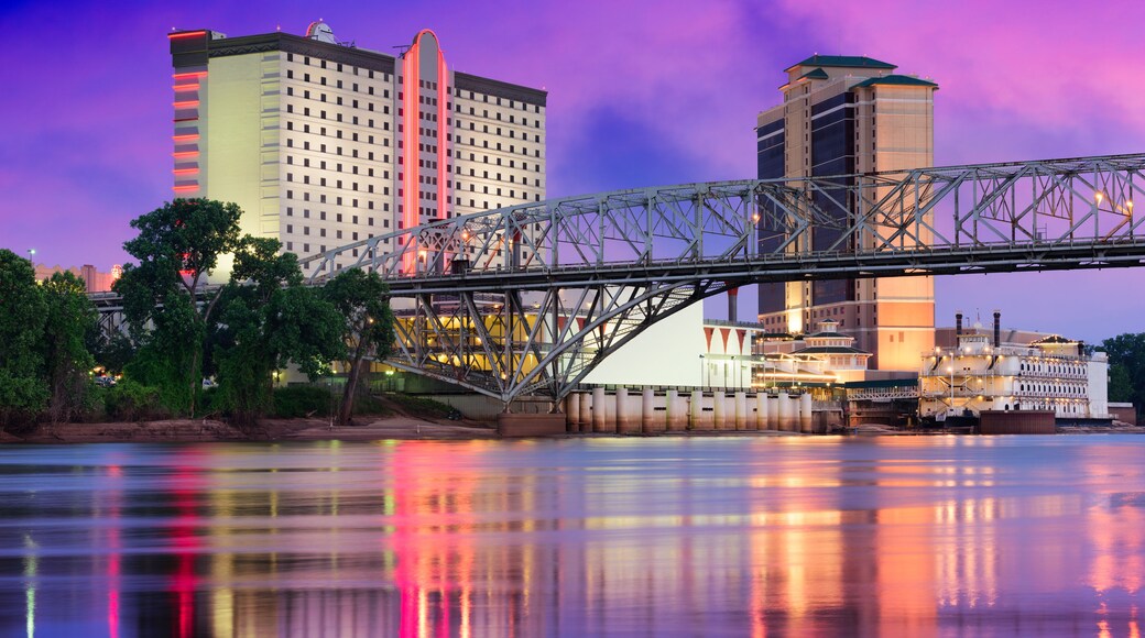 A bridge over a river with reflections of buildings and a city skyline at dusk.