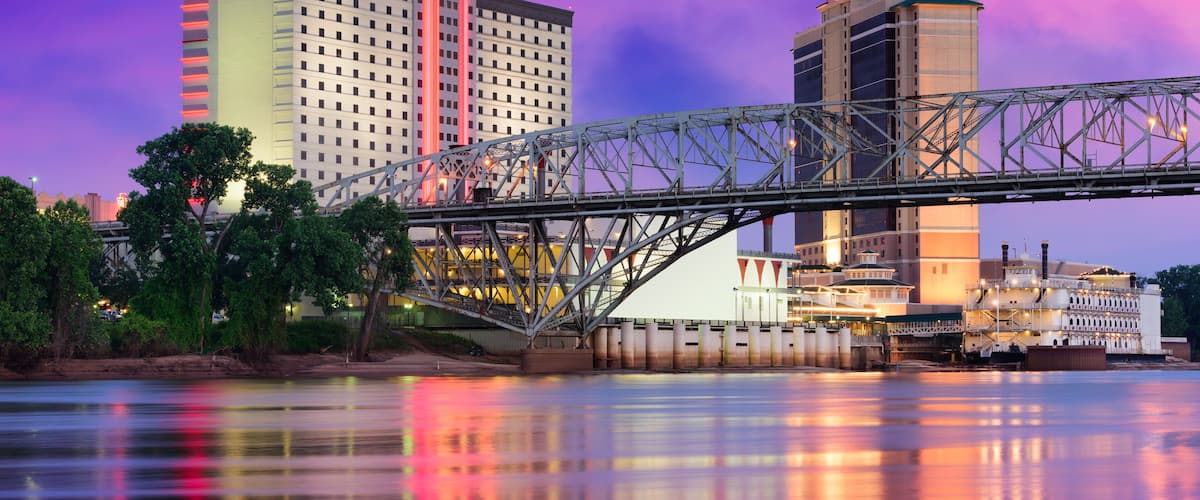 A bridge over a river with reflections of buildings and a city skyline at dusk.