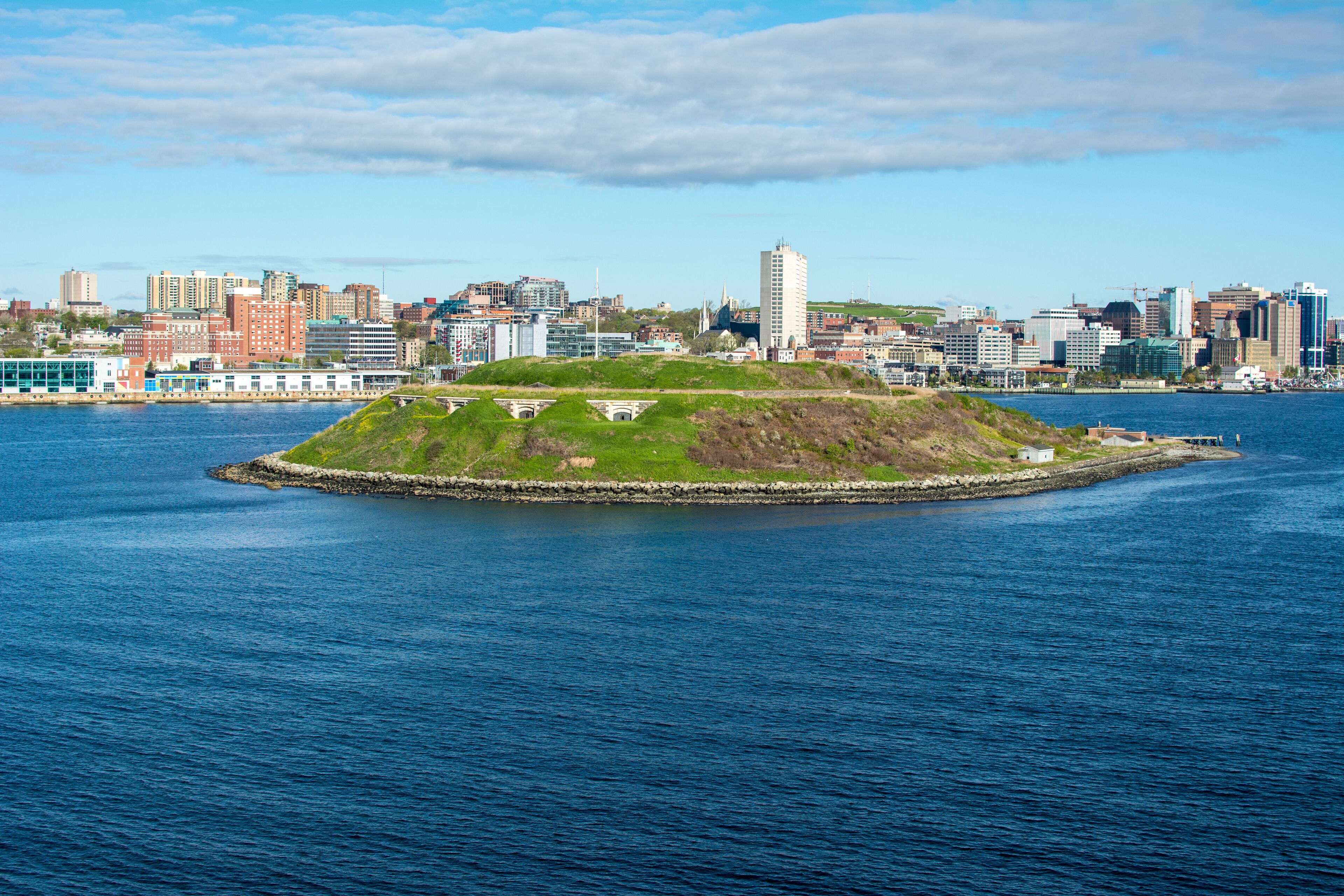 A coastal cityscape with a prominent hill with buildings on top, a large body of water, and a distant skyline of various buildings.