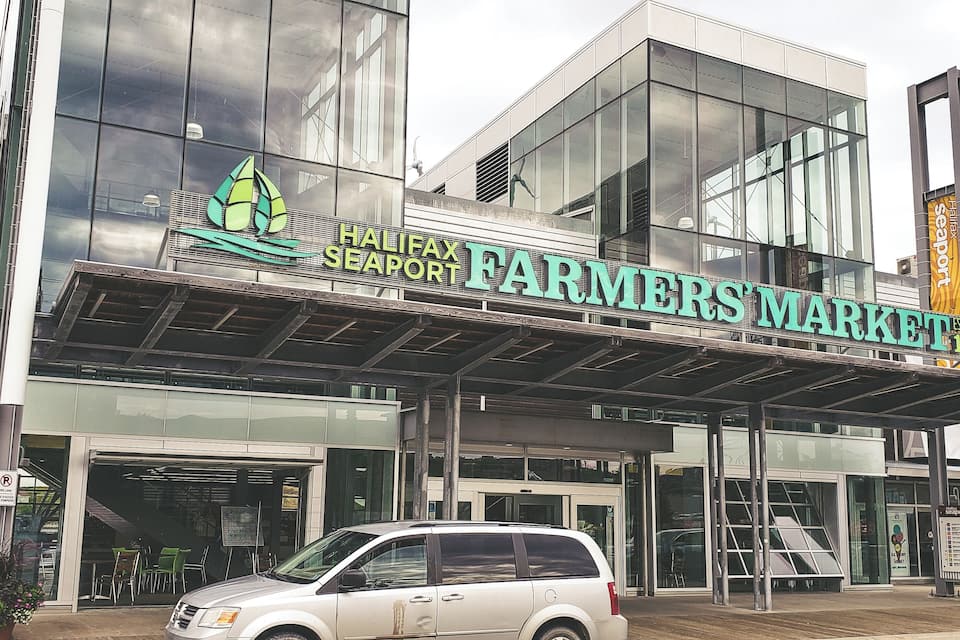 Fasaden på Halifax Seaport Farmers' Market, med en modern glasfasad, och en vit SUV parkerad framför.