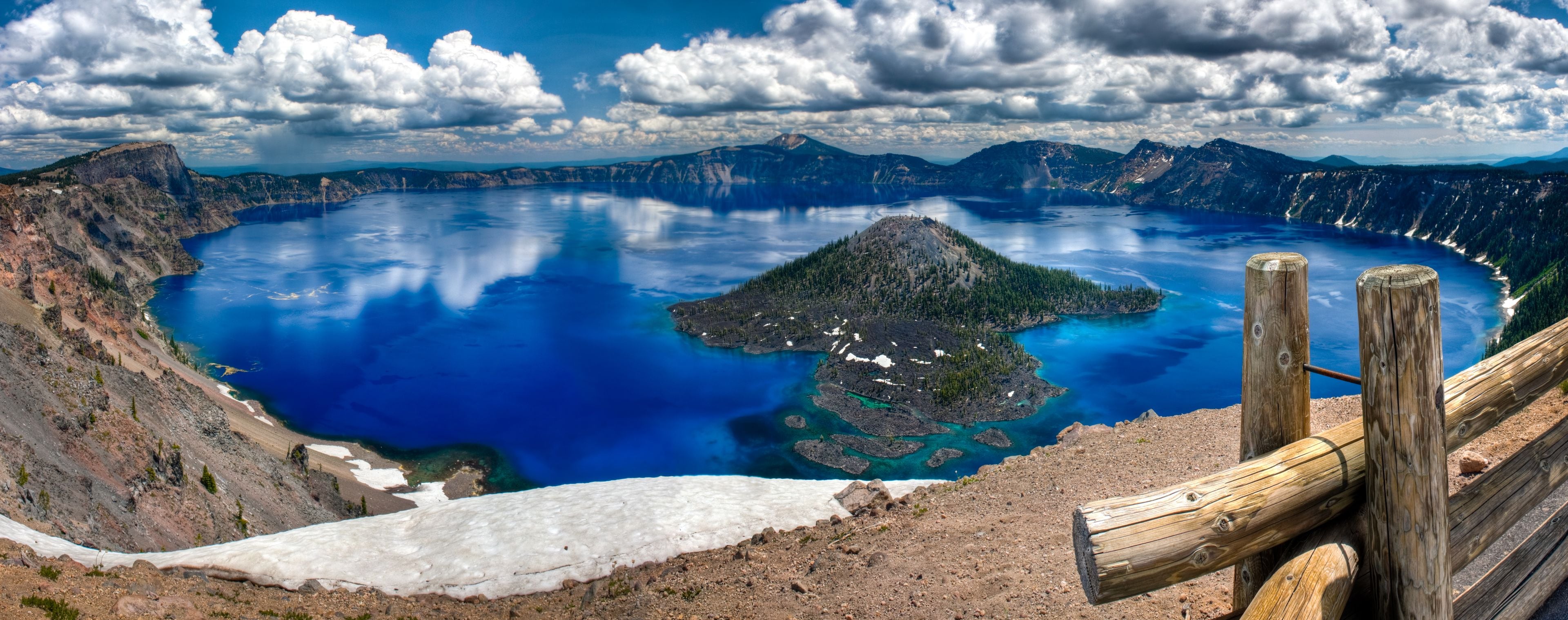 A large lake surrounded by mountains, a small island in the center, and a wooden fence in the foreground.