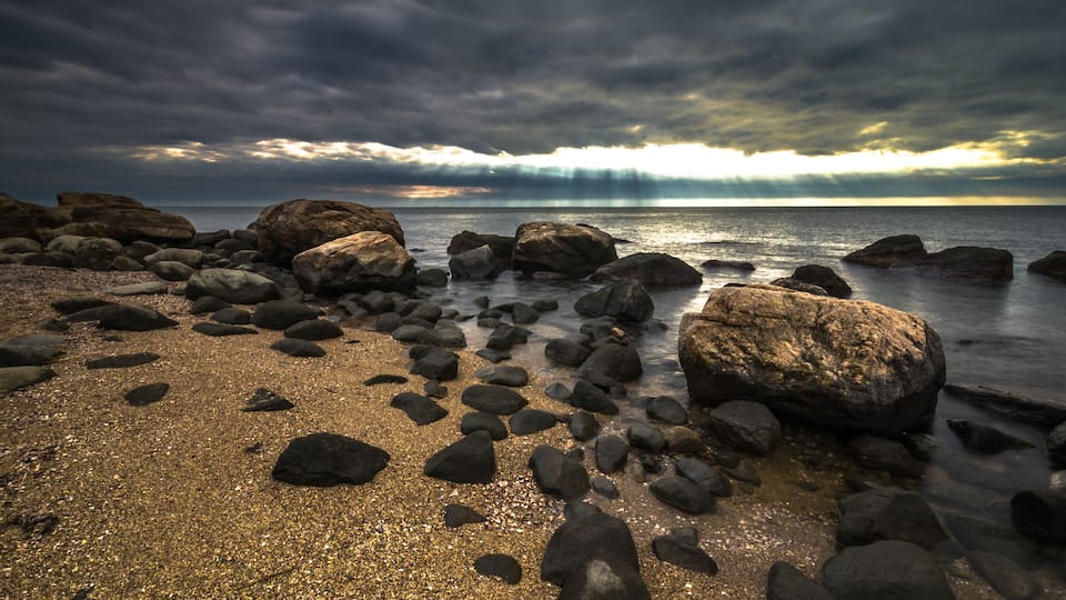 A rocky shoreline with large boulders and a calm sea under a cloudy sky.