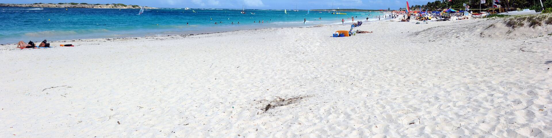 Una playa arenosa con agua azul cristalina, algunas embarcaciones y sombrillas en la orilla.