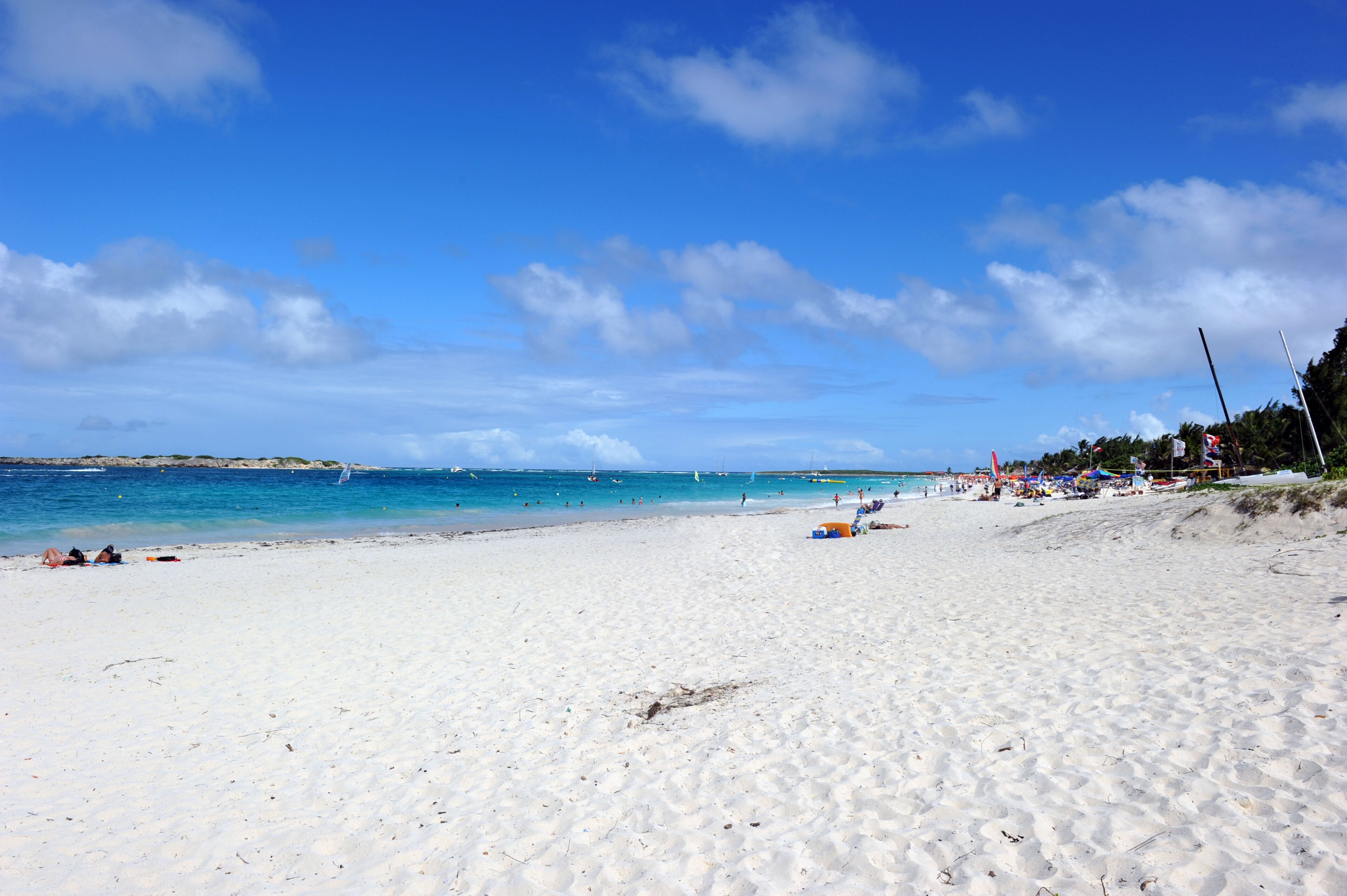A beach with white sand, clear blue water, and a few boats in the distance.