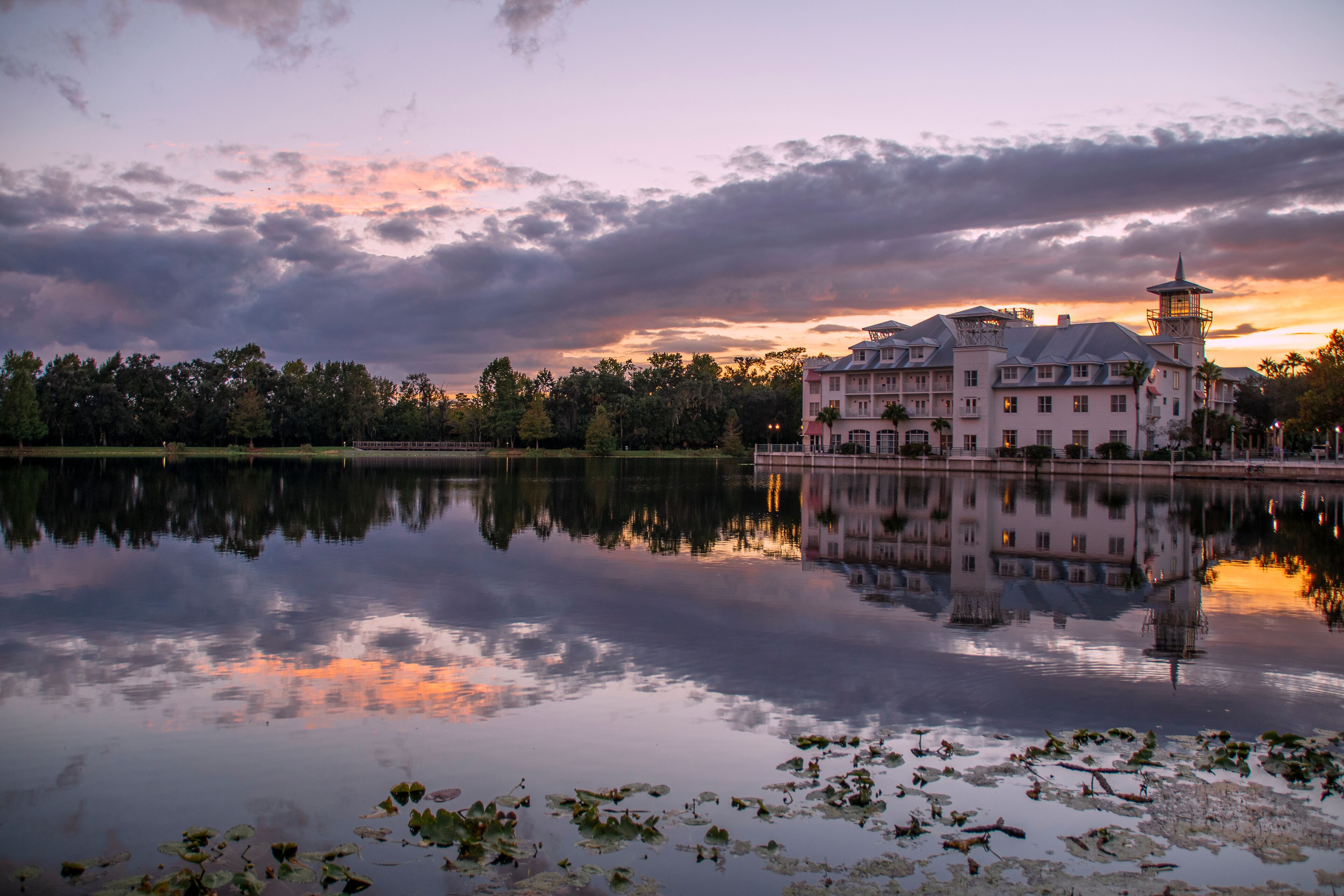 A hotel in Celebration (Orlando, Kissimmee), Florida reflects at the edge of a small lake during sunset with interesting cloud formations above.