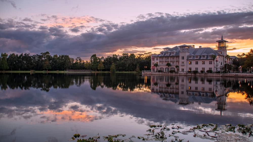 A large, historic building with a tower, reflecting on a calm lake with lily pads, surrounded by trees and a sunset sky.