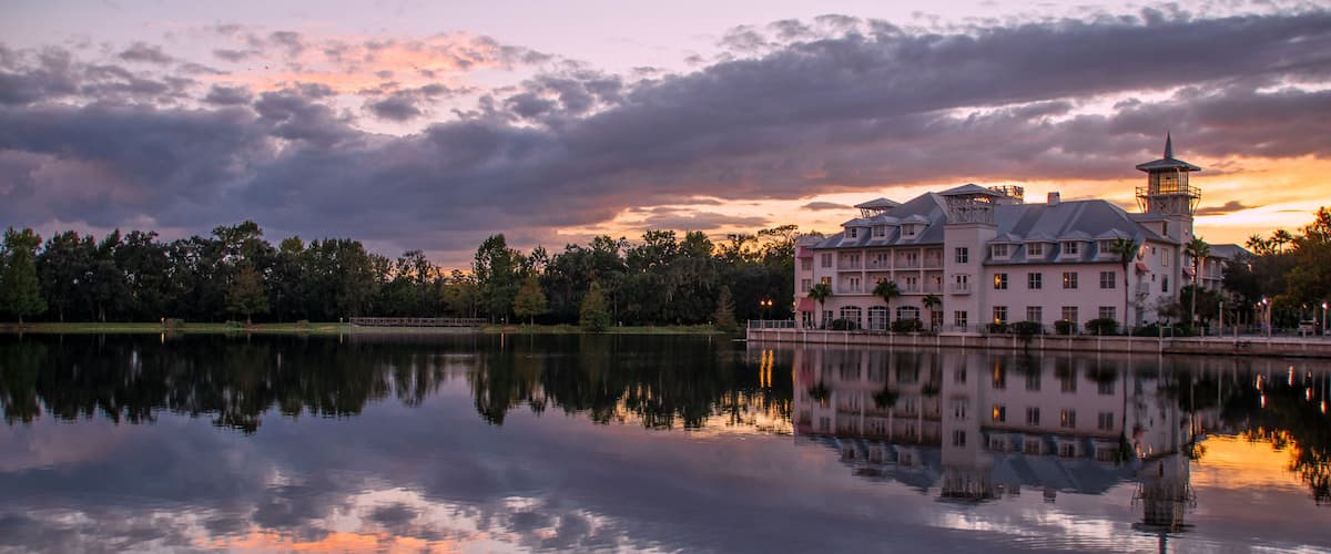 Un grand bâtiment historique, doté d’une tour, qui se reflète sur un lac paisible parsemé de nénuphars, entouré d’arbres et illuminé par le ciel au coucher du soleil.