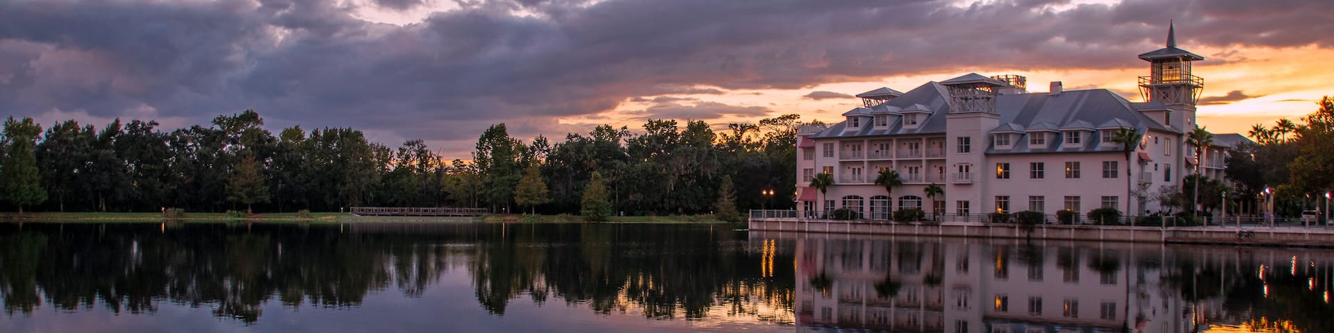 A large, historic building with a tower, reflecting on a calm lake with lily pads, surrounded by trees and a sunset sky.