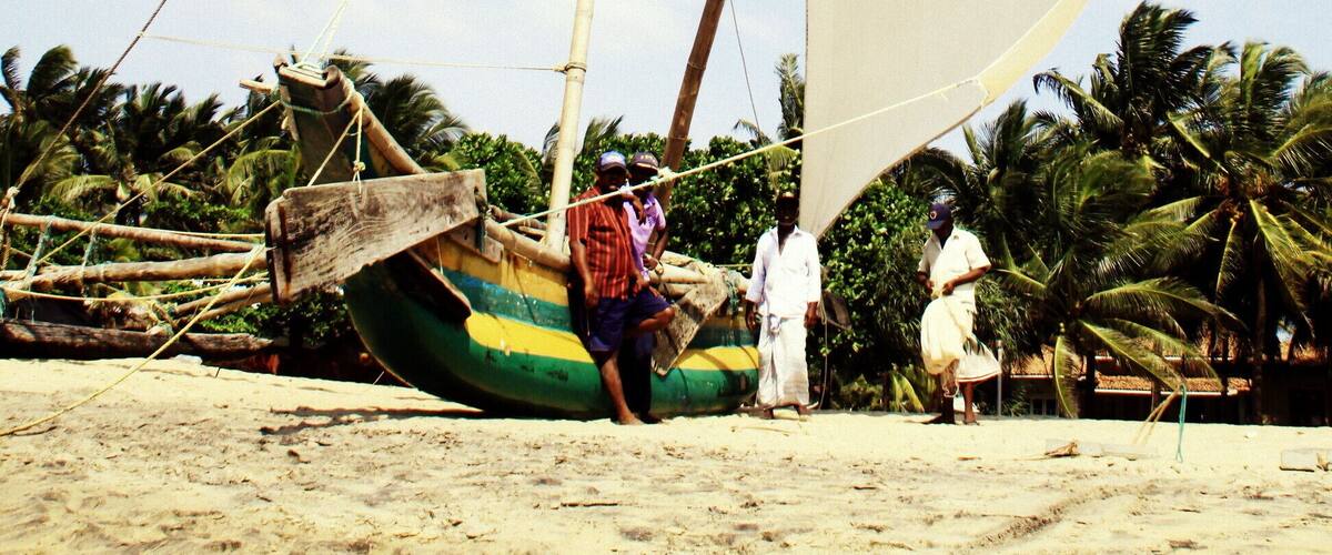 A traditional sailboat on a sandy beach with palm trees in the background.