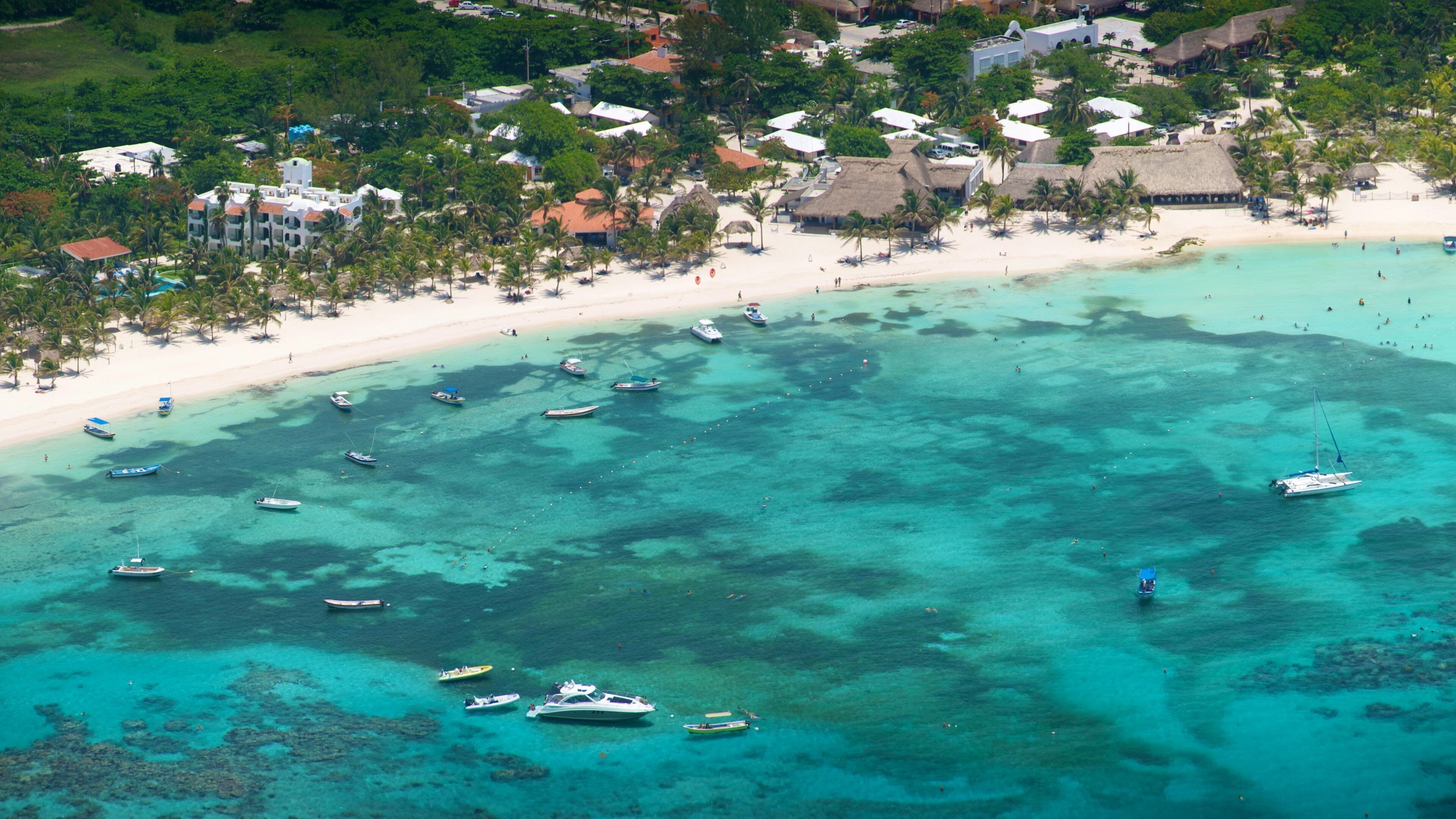 A tropical beach with clear turquoise waters, numerous boats, and a resort area with thatched-roof buildings.