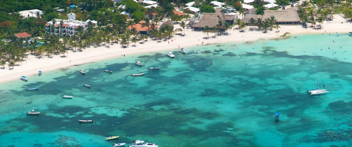 A tropical beach with clear turquoise waters, numerous boats, and a resort area with thatched-roof buildings.