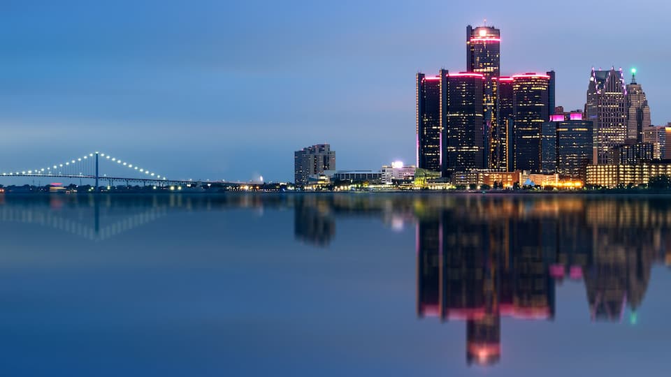 A city skyline with illuminated buildings and a bridge reflected in the water.