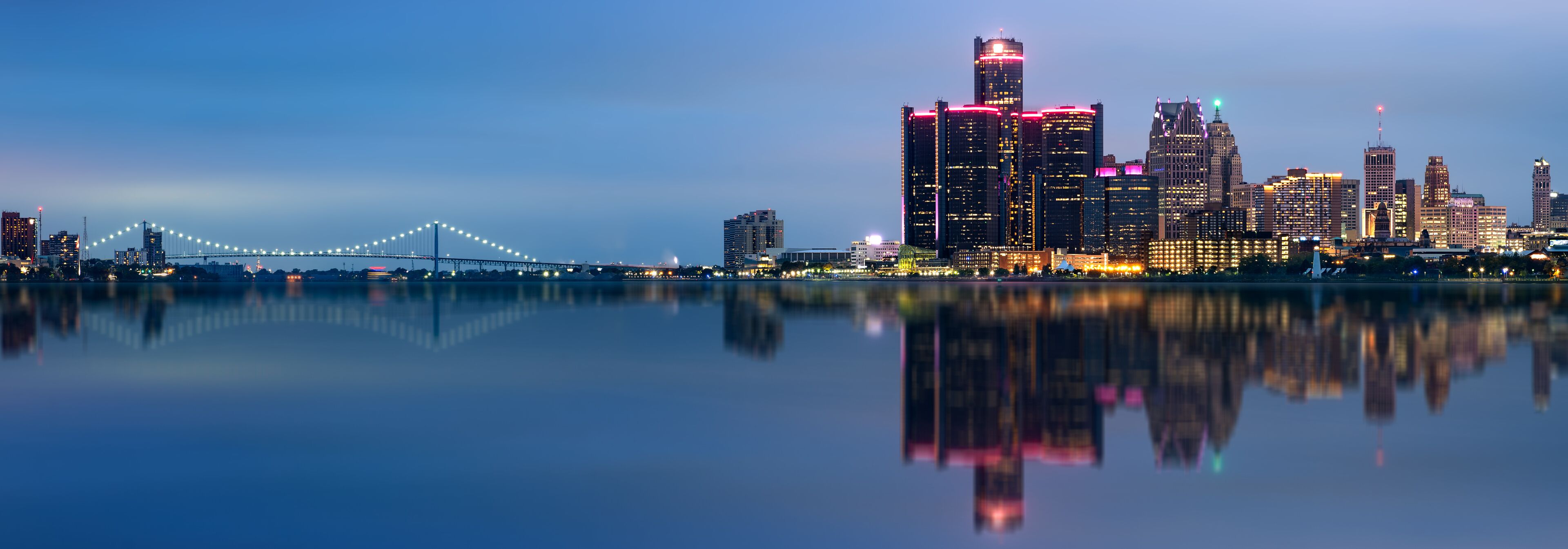 A city skyline with illuminated buildings and a bridge reflected in the water.