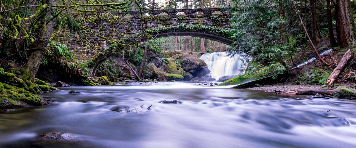 Un pont de pierre enjambant une rivière aux eaux vives, entouré d’une végétation luxuriante et d’une cascade.
