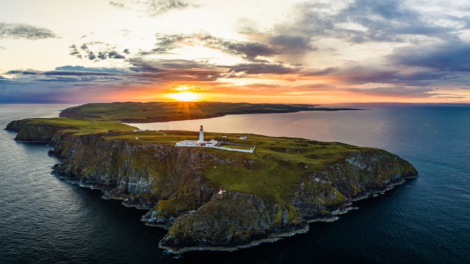 A coastal landscape with a lighthouse on a small island, surrounded by water and a sunset in the background.