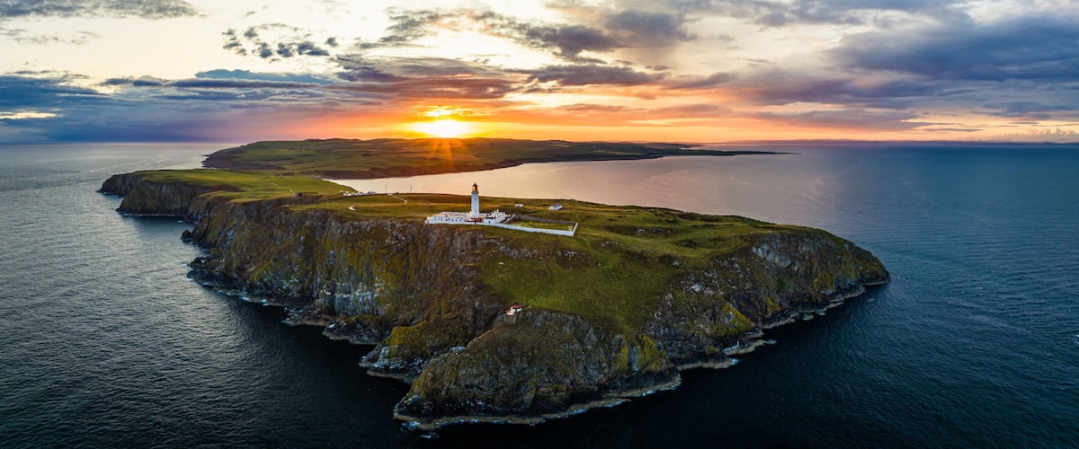 A coastal landscape with a lighthouse on a small island, surrounded by water and a sunset in the background.