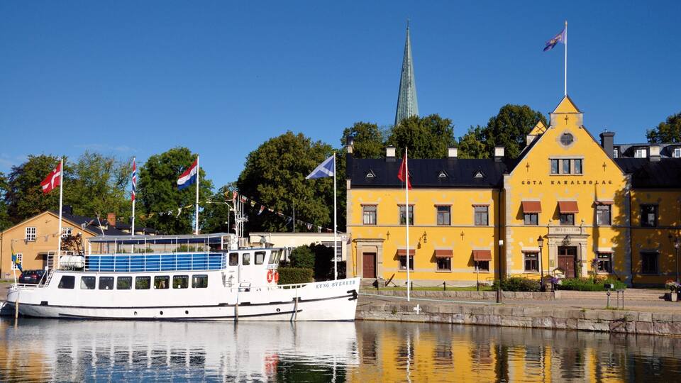 A river with a boat docked at a pier, a yellow building with flags, and a church spire in the background.