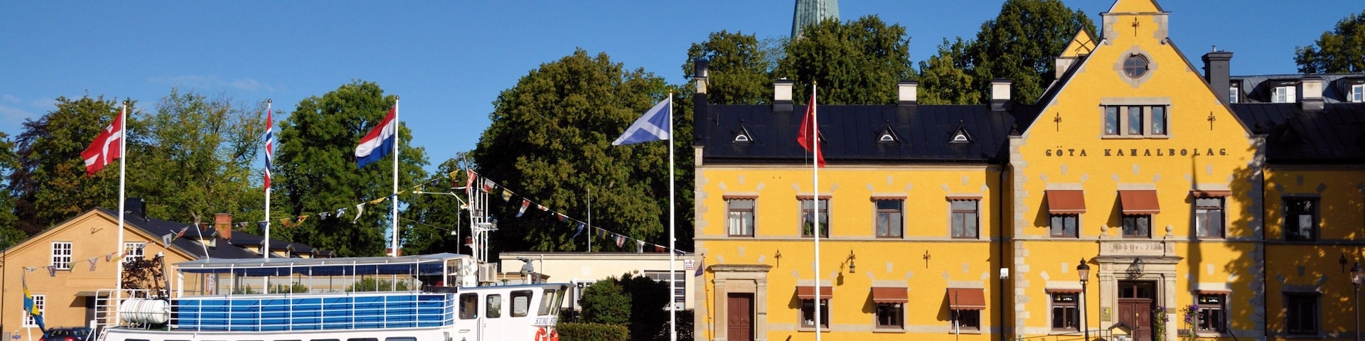 A river with a boat docked at a pier, a yellow building with flags, and a church spire in the background.