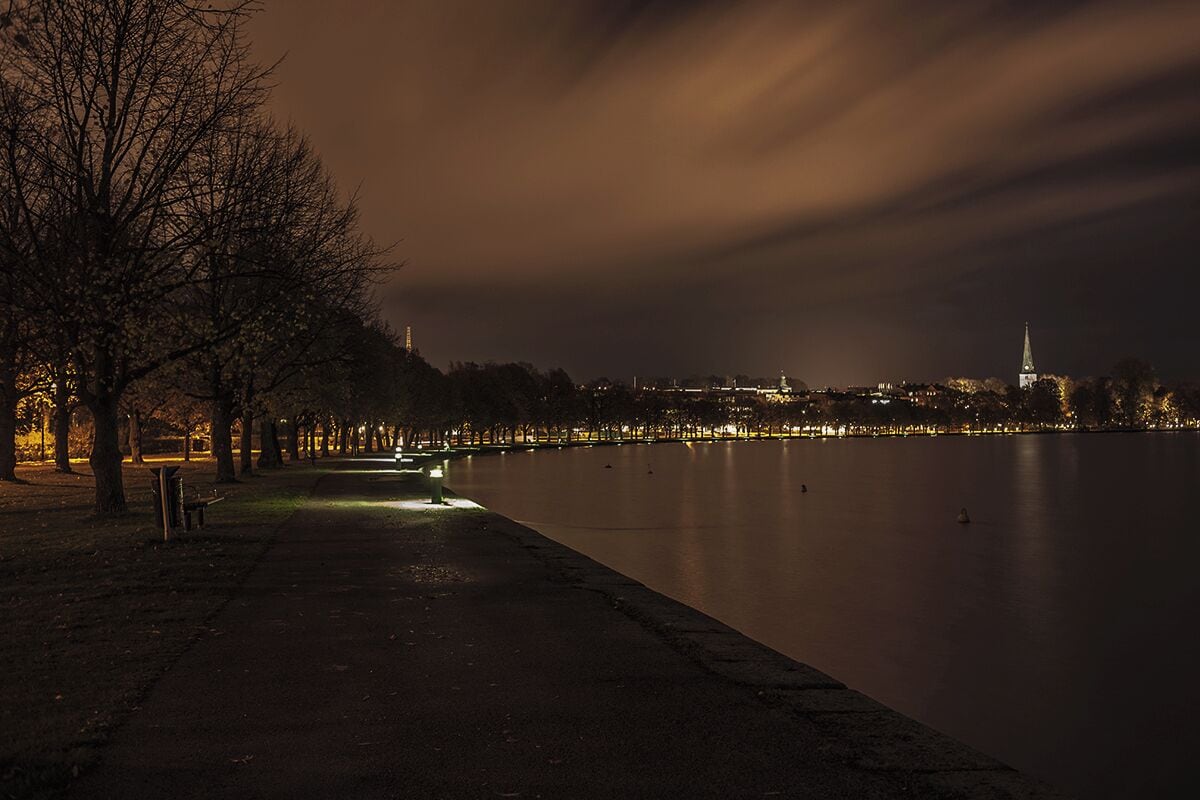Vista nocturna de un lago tranquilo con un parque en una orilla, un horizonte urbano distante y una torre destacada.