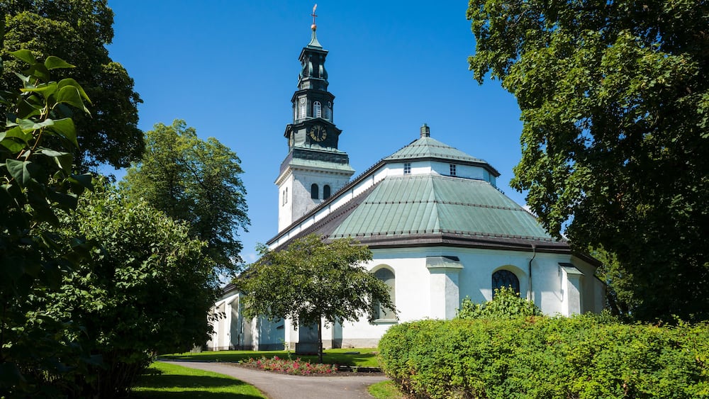 Köping church, white paint facade, copper-covered gable roof, Baroque style spire on copper-clad tower with clock, Old religious building surrounded by green garden, Sweden