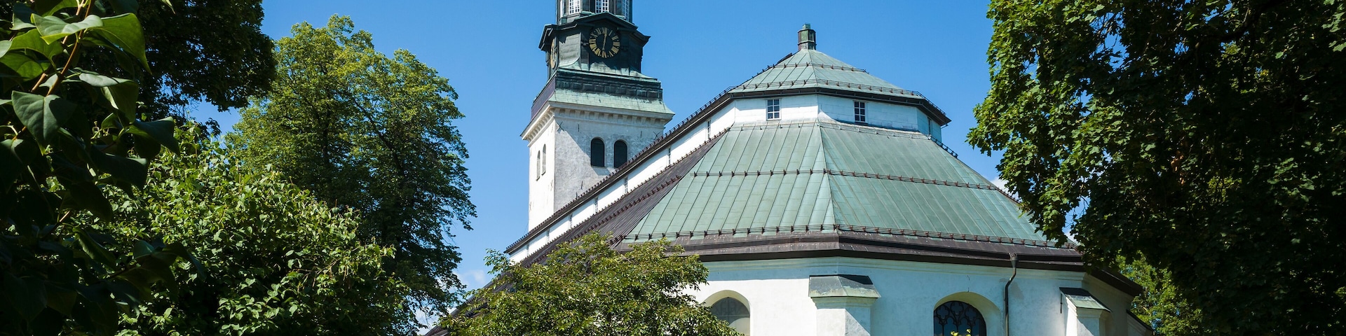 Köping church, white paint facade, copper-covered gable roof, Baroque style spire on copper-clad tower with clock, Old religious building surrounded by green garden, Sweden