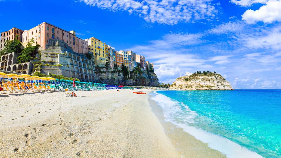 A beach with clear turquoise waters, a row of colorful umbrellas, and historic buildings on a cliff.
