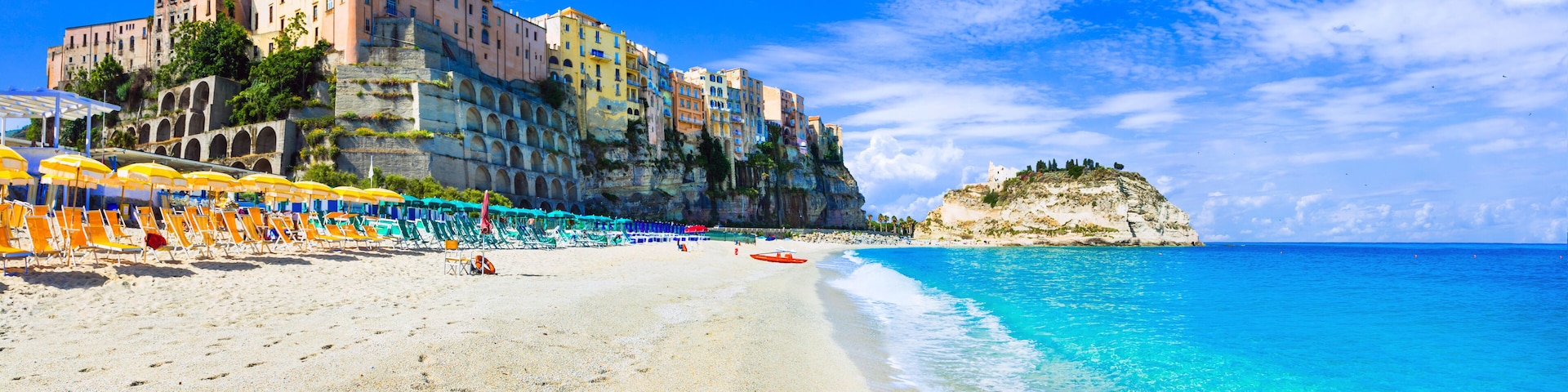Uma praia com águas turquesa cristalinas, uma fileira de guarda-sóis coloridos e edifícios históricos em um penhasco.
