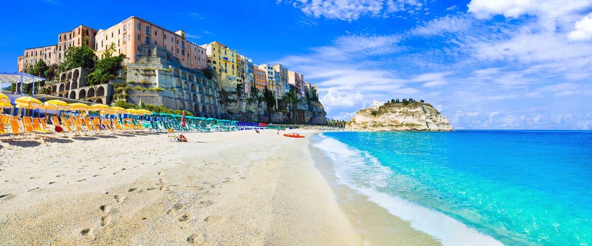 A beach with clear turquoise waters, a row of colorful umbrellas, and historic buildings on a cliff.