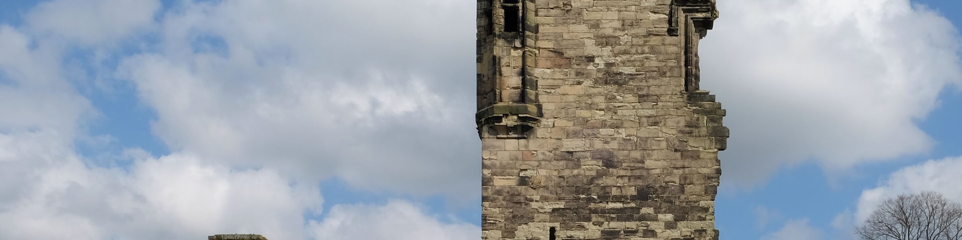 Una torre de piedra medieval con almenas, rodeada de campos verdes y árboles bajo un cielo parcialmente nublado.